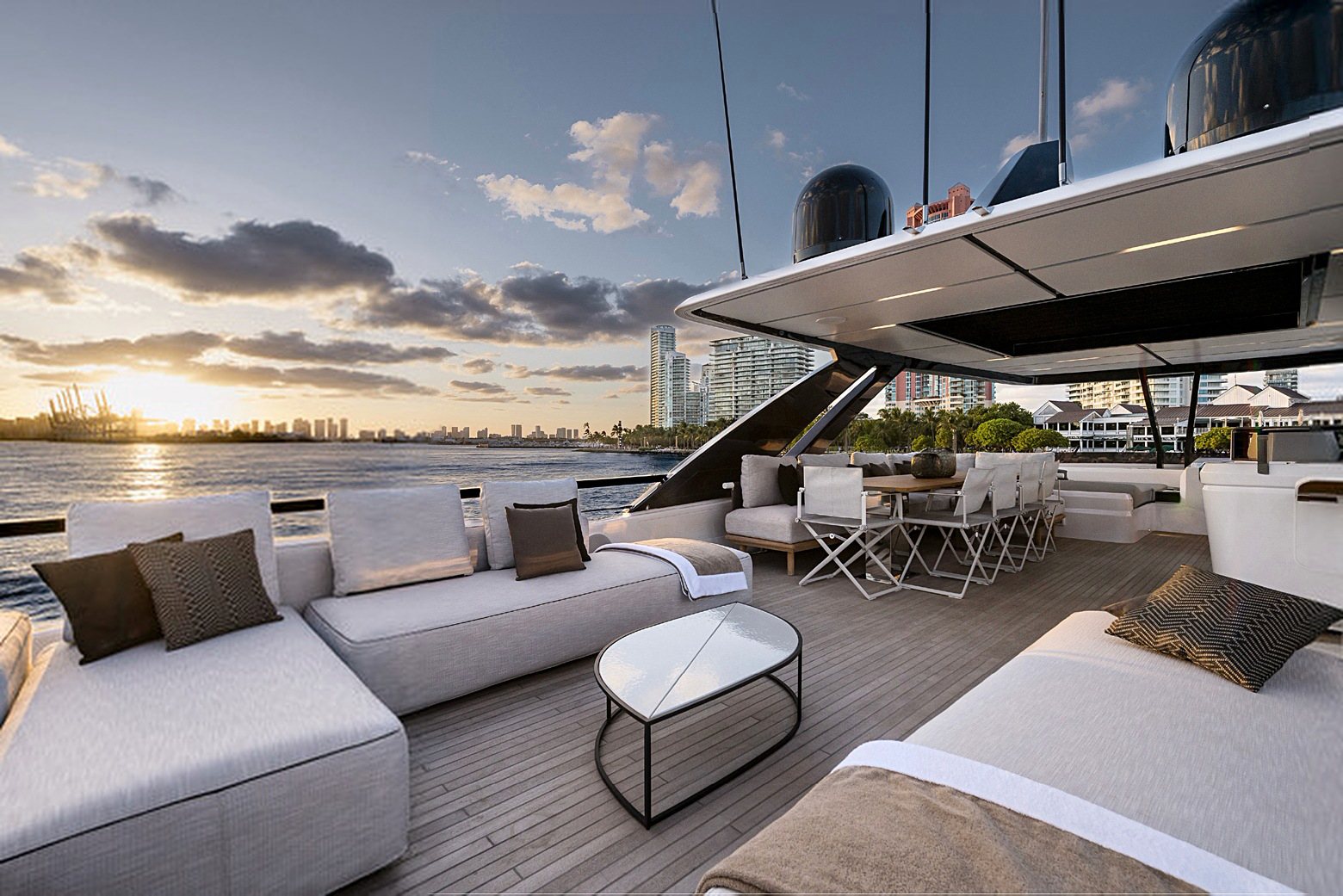 Aft deck at sunset: a long sunpad, a low table and a dining setting aft, with the city on the horizon.