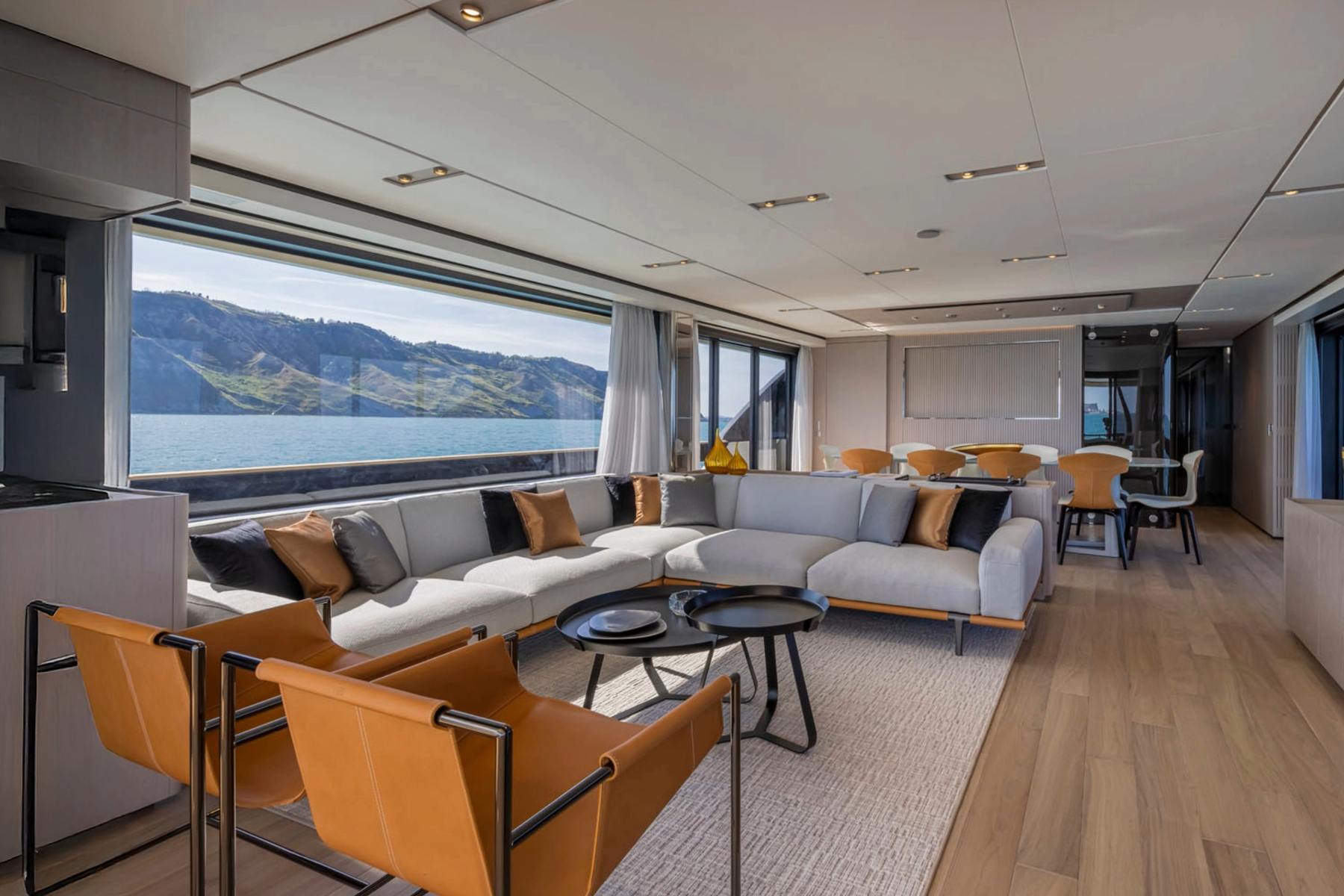 Main salon of a delivered yacht: a long pale sectional, two tan leather armchairs and a wide window onto the coast.